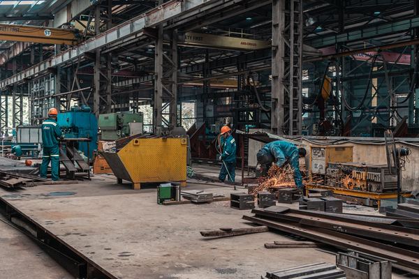 Workers in hard hats grind and cut steel inside a large metal workshop.