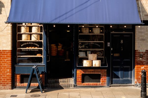 A small cheese shop with a navy awning and cheese wheels stacked in the windows.