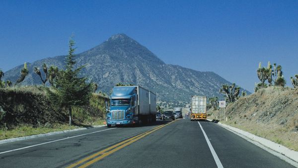 Freight trucks roll along a two-lane highway beneath a mountain.