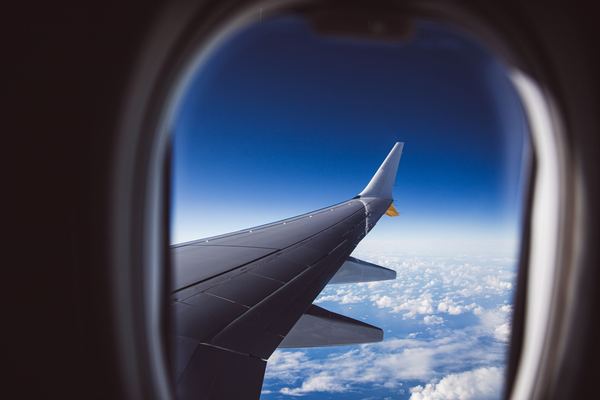 An airplane wing above the clouds, framed by a cabin window.